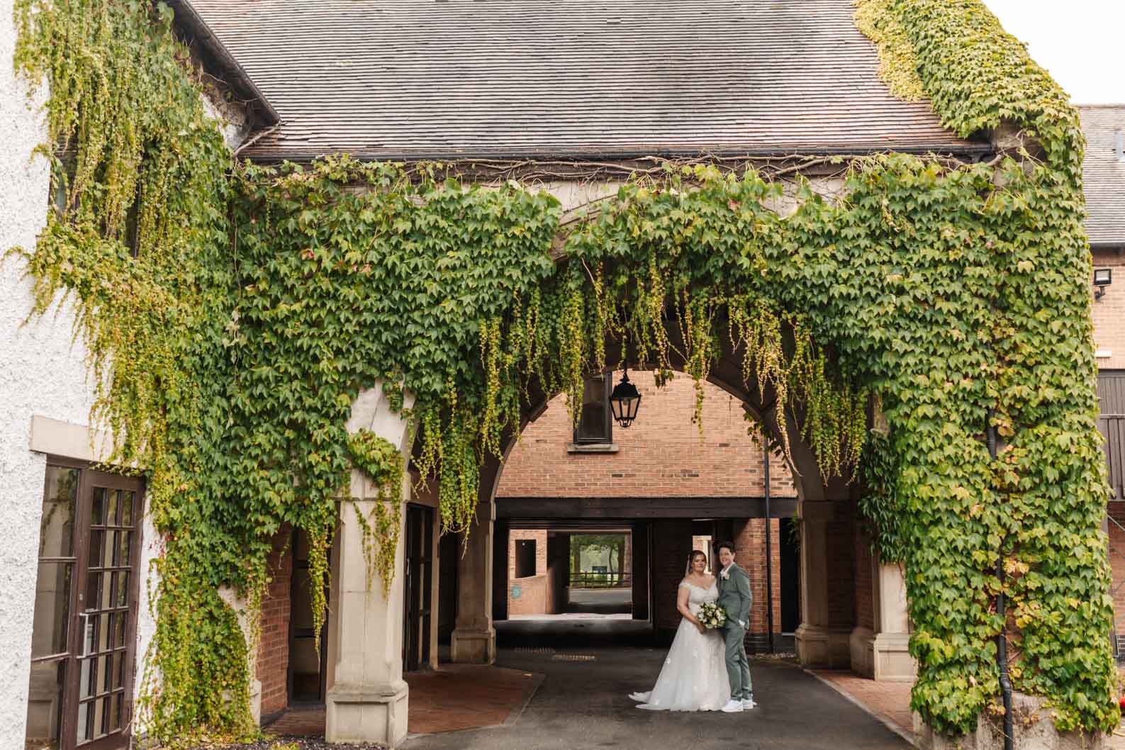 2 Brides standing under a covered archway during light rain at a West Midlands wedding venue