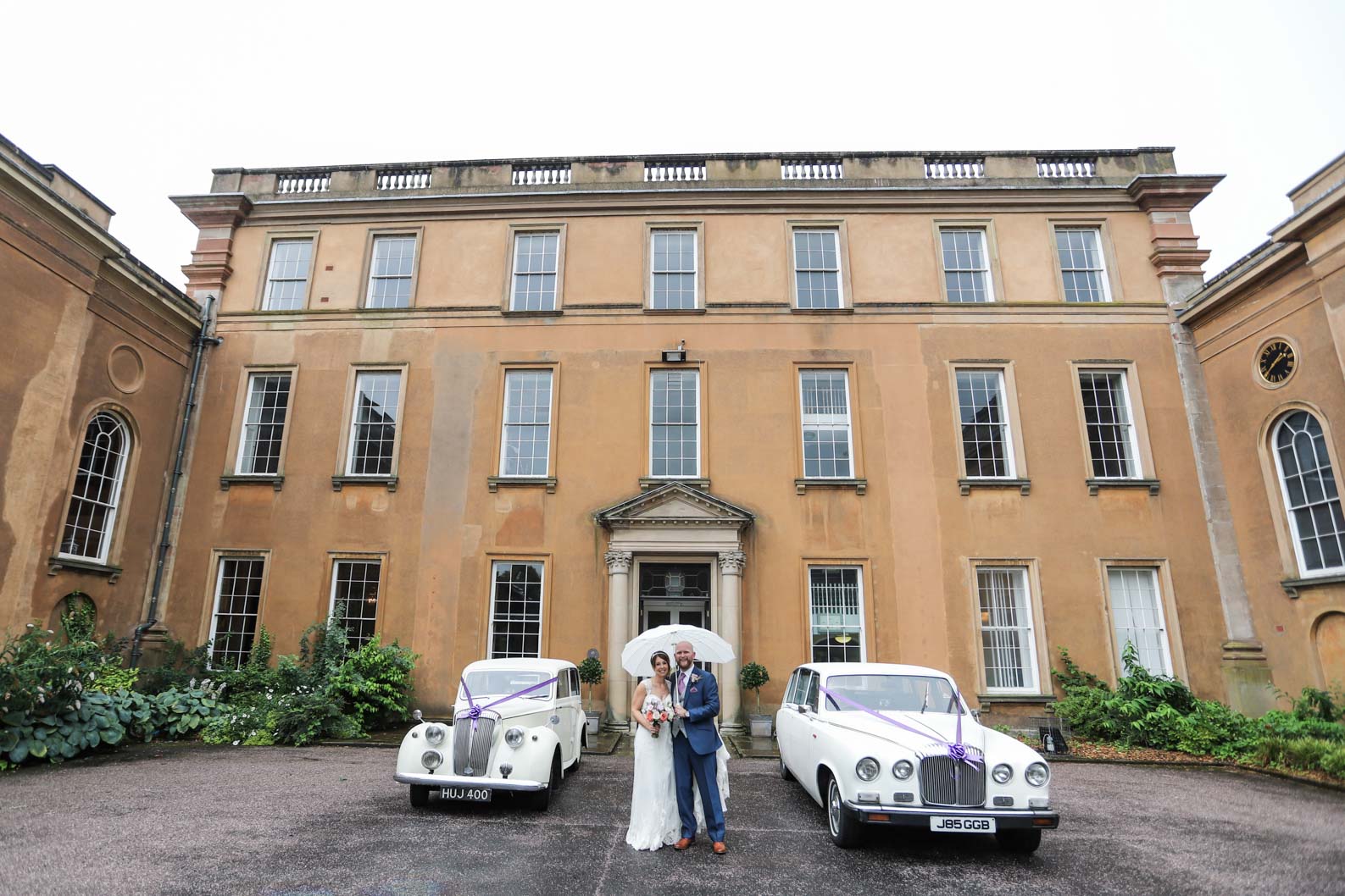Bride and groom posing with white umbrellas in front of white cars outside Himley Hall during a rainy West Midlands wedding