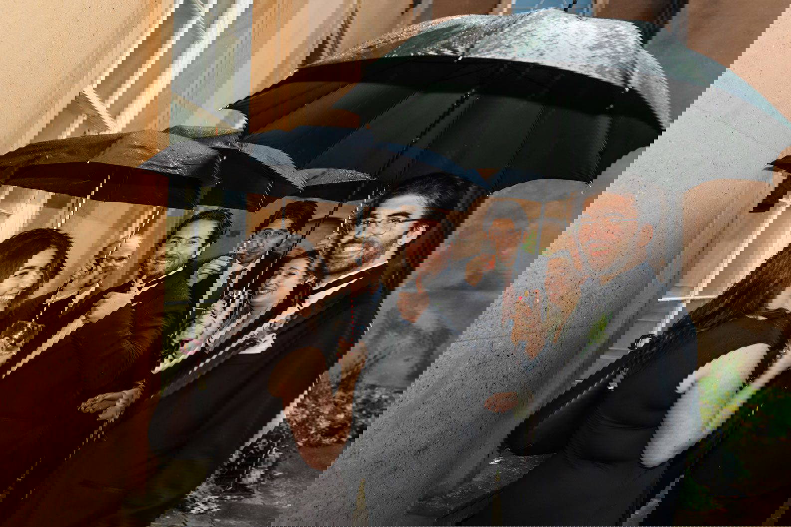 Group of friends smiling and huddled together under black umbrellas outside Himley Hall during a rainy West Midlands wedding