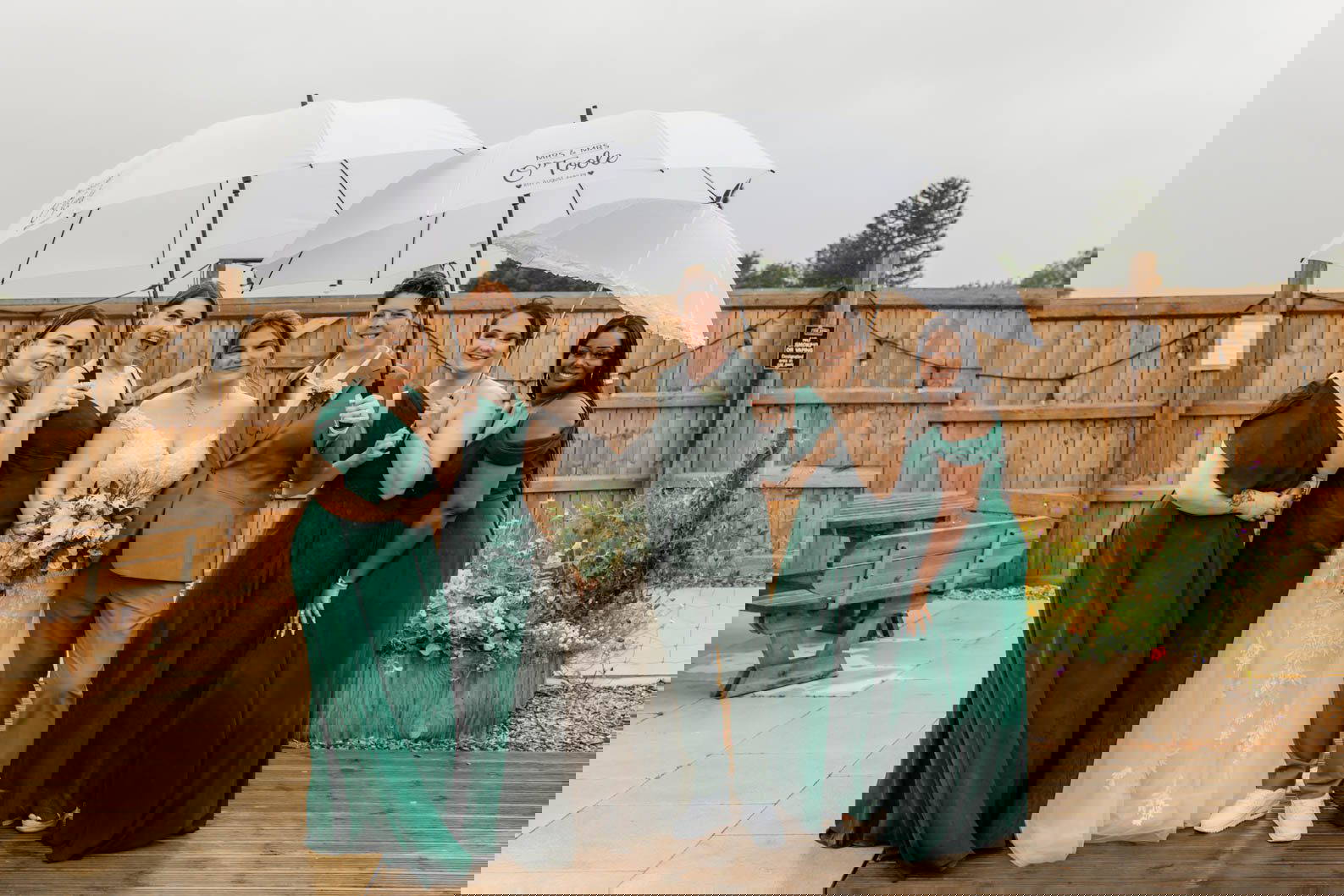 2 brides with their bridesmaids posing for a group photo in the rain with umbrellas